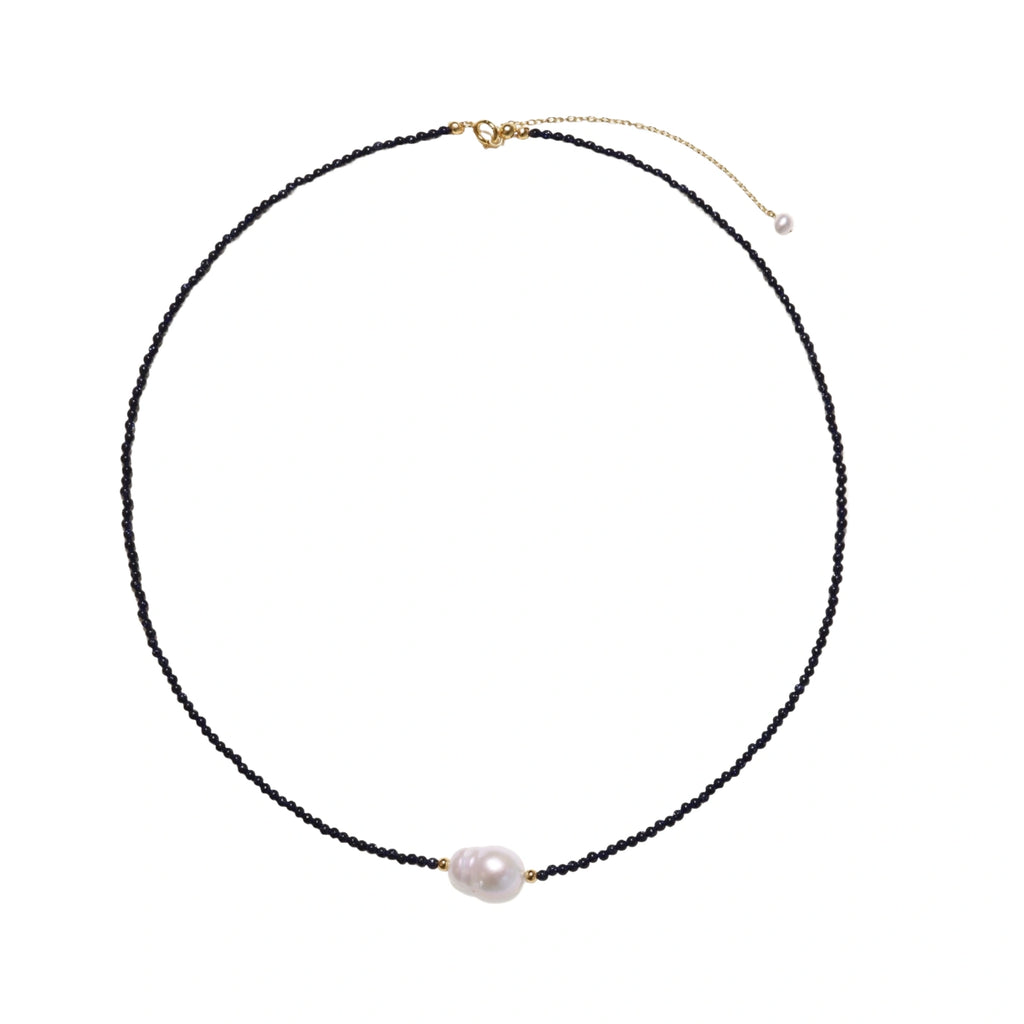 Delicate necklace with black blue sandstone beads and baroque pearl center on white background with gold accents.