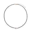Delicate necklace with black blue sandstone beads and baroque pearl center on white background with gold accents.