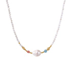 Delicate white beaded necklace with baroque pearl center, gold accents, and coral and aqua gemstones on cream background.