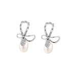 Silver bow-shaped stud earrings with baguette crystals and dangling white freshwater pearls on cream background