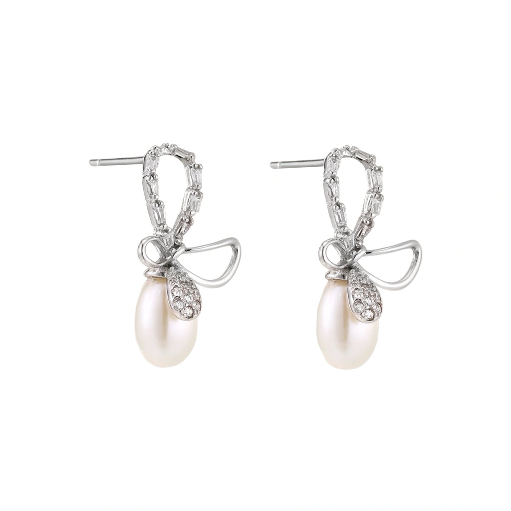 Silver bow-shaped stud earrings with baguette crystals and dangling white freshwater pearls on cream background