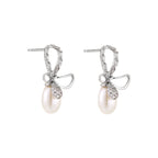 Silver bow-shaped stud earrings with baguette crystals and dangling white freshwater pearls on cream background