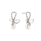 Silver bow-shaped stud earrings with baguette crystals and dangling white freshwater pearls on cream background